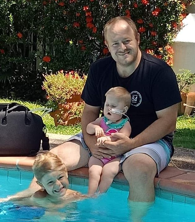 Brett beside a pool holding his daughter Florence, with his older child in the water