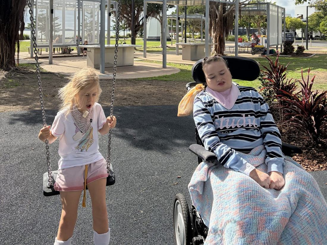 Florence on a swing at a Gladstone park on a sunny afternoon, with Shontae in her wheelchair beside her