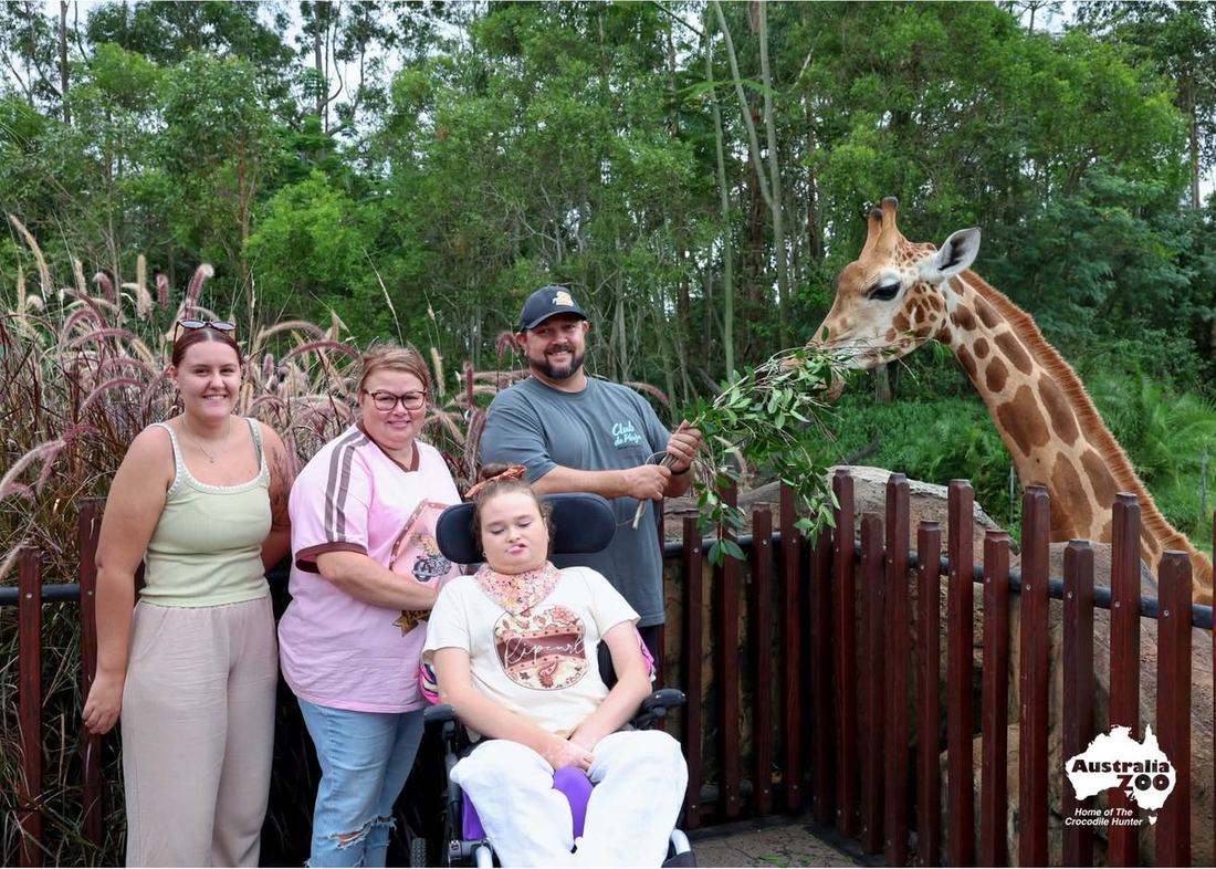 Shontae in her wheelchair at Australia Zoo with her family, watching a giraffe feed from branches held out over the fence