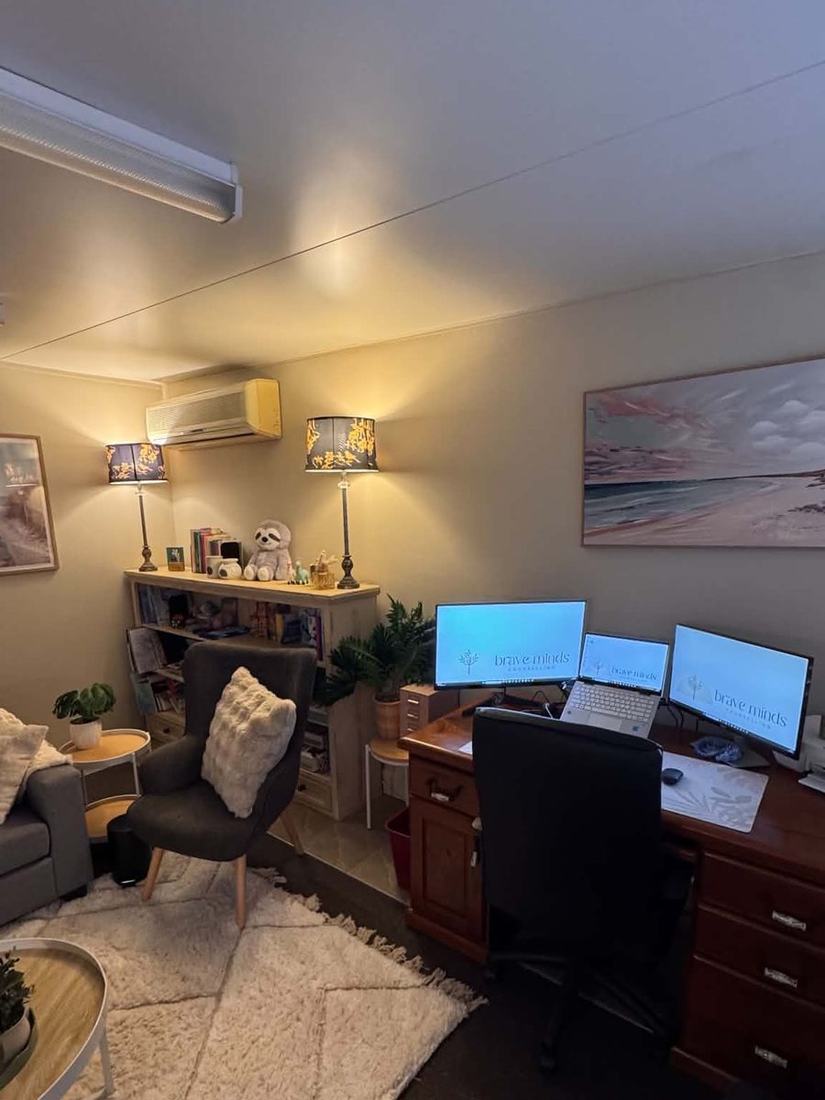The desk in the corner of the counselling room, with Brave Minds Counselling on the screens and lamps either side of a bookshelf