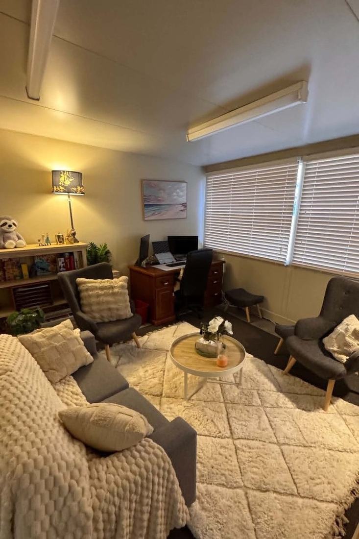 The Brave Minds counselling room: two armchairs and a couch around a low table with flowers, a lamp, a bookshelf with books and a soft toy, and a window with blinds
