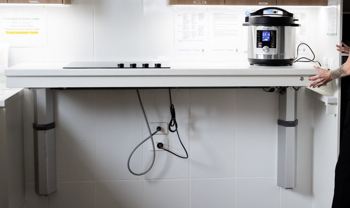 A kitchen bench on motorised legs with an induction cooktop set into it, raised to standing height, with a hand on the height control at one end